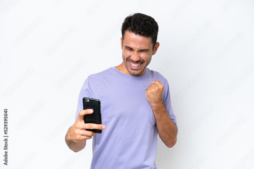 Young handsome man over isolated white background using mobile phone and doing victory gesture Young handsome man over isolated white background using mobile phone and doing victory gesture