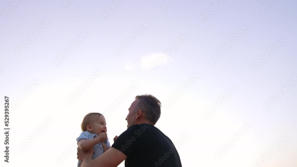 Mid-aged man tosses little baby boy in the air. Adorable kid is excited and laughing cheerfully. Low angle view. Sky at backdrop. Mid-aged man tosses little baby boy in the air. Adorable kid is excited and laughing cheerfully. Low angle view. Sky at backdrop.
