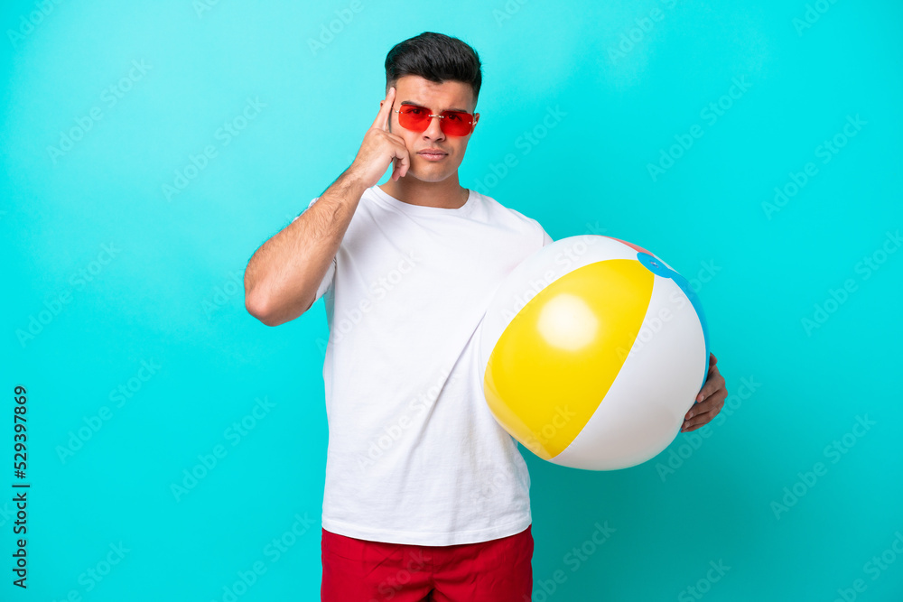 Young caucasian man holding a beach ball isolated on blue background thinking an idea