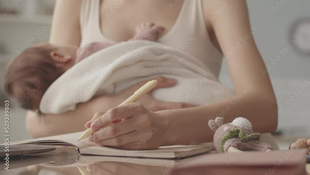 Cropped slowmo of unrecognizable young woman with newborn baby in her arms taking notes in copybook while studying or working from home Cropped slowmo of unrecognizable young woman with newborn baby in her arms taking notes in copybook while studying or working from home