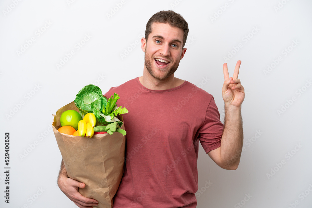 Young caucasian man holding a grocery shopping bag isolated on white background smiling and showing victory sign Young caucasian man holding a grocery shopping bag isolated on white background smiling and showing victory sign