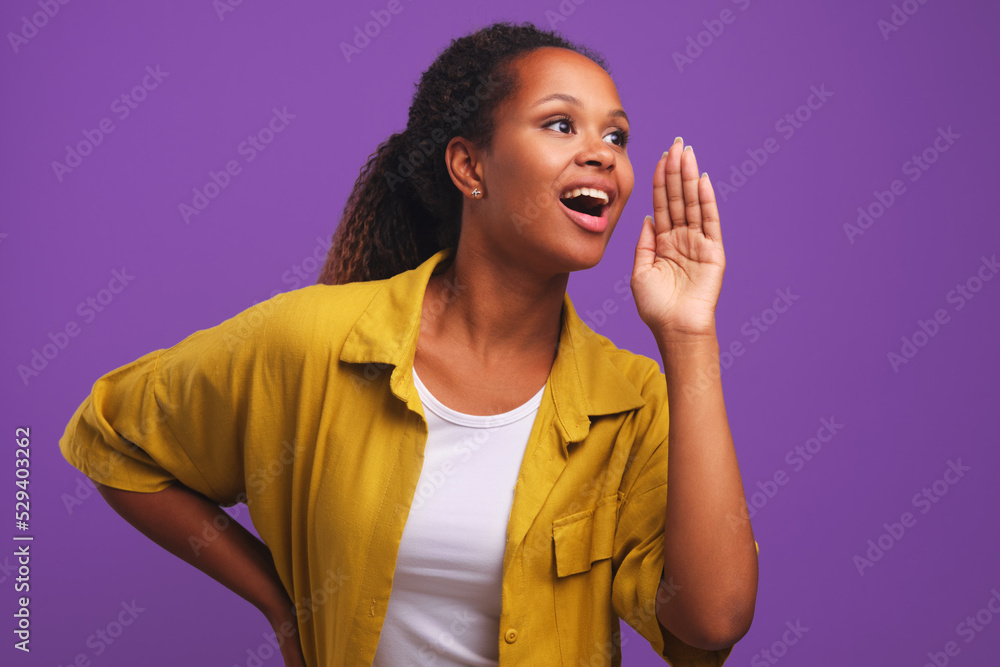 Excited young dark haired African American woman shouting to announce good shopping discounts and delight shoppers best price dressed in casual clothes stands on purple studio background