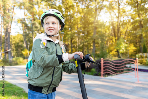 Profile view portrait of cute blond little caucasian school girl wear helmet enjoy having fun riding electric scooter city street park outdoors on sunny day. Healthy sport children activities outside photo