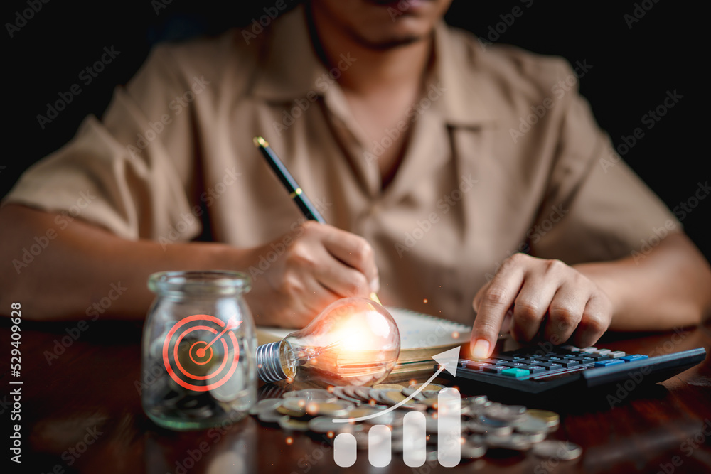 Man using calculator to calculate and money stack, coin in a jar, lightbulb on desk with note book. idea saving energy and accounting finance concept. Man using calculator to calculate and money stack, coin in a jar, lightbulb on desk with note book. idea saving energy and accounting finance concept.