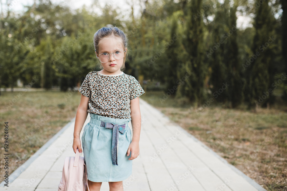 Funny cute child girl 4 5 year old wear glasses and casual clothes holding backpack in park over nature background. Childhood. Back to school. Stock Adobe Stock