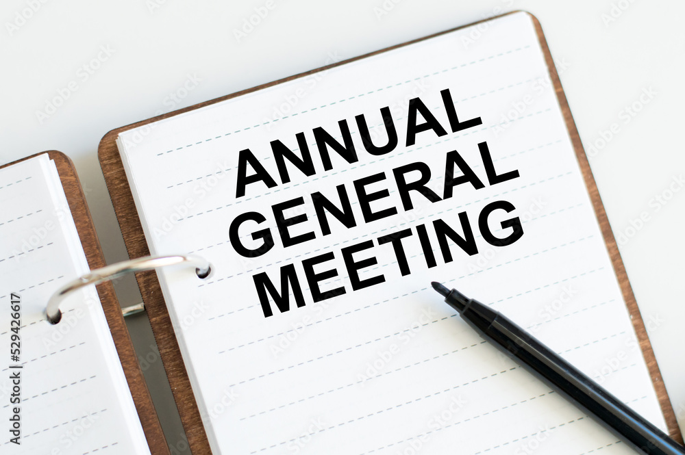 On a light background, a white notebook with are words AGM Annual General Meeting and a pen. On a light background, a white notebook with are words AGM Annual General Meeting and a pen.