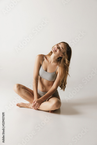 Young adorable smiling woman in lingerie sitting on floor isolated over gray background. Wellness, wellbeing, fitness and aesthetic cosmetology concept.