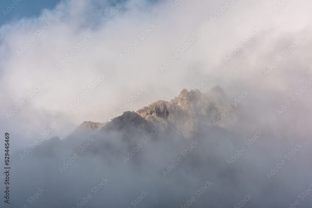 Lovely scenery with big mountain peak in thick clouds in sunlight. Sunlit high pinnacle in gantly cloudy sky. Large mountain top in clearance of dense fog. Beautiful rocks in low clouds in sunrise.