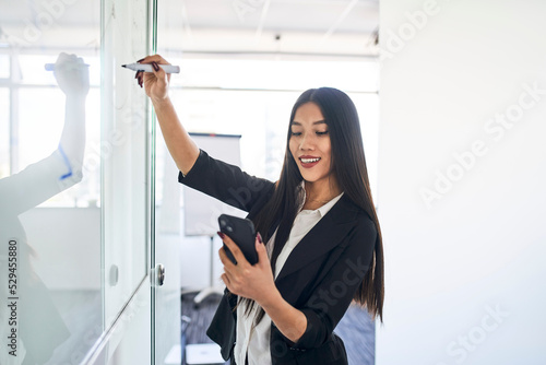 Young Asian businesswoman using her smartphone while writing on whiteboard in office