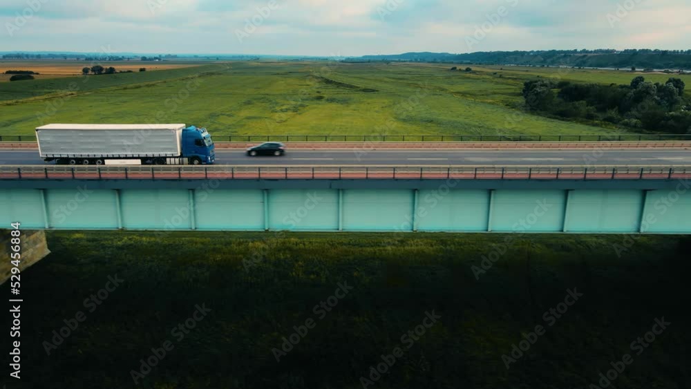 Blue truck with grey trailer fast driving on highway by bridge and delivers goods to customers. Concept of optimizing cargo transportation business Blue truck with grey trailer fast driving on highway by bridge and delivers goods to customers. Concept of optimizing cargo transportation business
