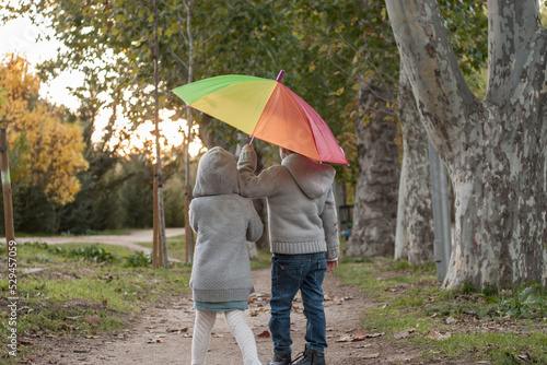 dos gemelos paseando por un parque en otoño con un paraguas aco iris	 photo