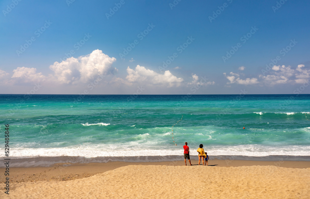 Beautiful calm seascape with horizon line and clouds in blue sky, light surf and strip of golden sandy beach and people with view from back.