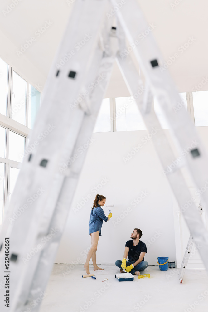 Young couple in love, the newlyweds are making repairs in an empty room, painting white walls, updating the house. A happy man and woman took out a mortgage and moved into a new house. Place for text 