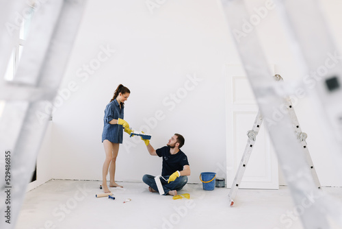 Young couple in love, the newlyweds are making repairs in an empty room, painting white walls, updating the house. A happy man and woman took out a mortgage and moved into a new house. Place for text 