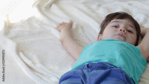 kid lies on bed morning sunshine day.child moving hands and legs similar of doing snow angel.little boy feeling safe secure cozy comfort at home with family mother.ivory blanket blue clothes photo