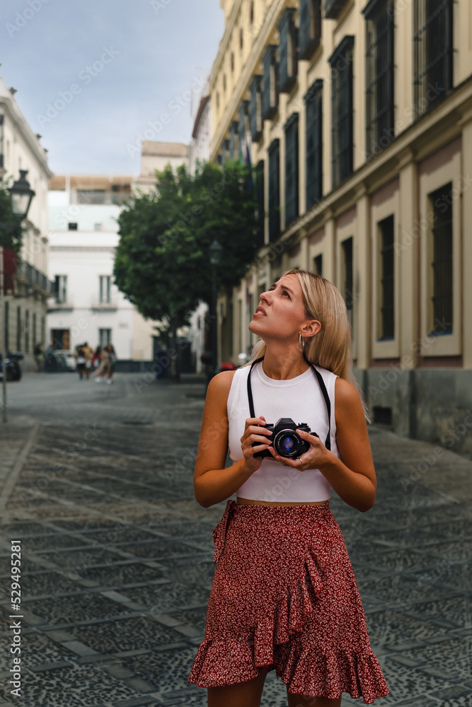 Portrait of a young blonde woman tourist in Seville looking for a photo with her camera. Concept of tourism, photography and monumental architecture.
