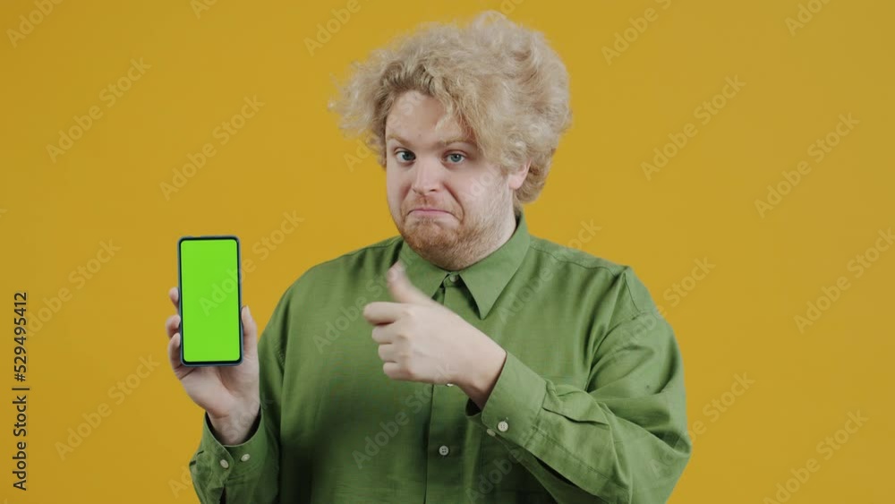 Portrait of satisfied man pointing at green chroma key smartphone screen and showing thumbs-up gesture standing on yellow color background Portrait of satisfied man pointing at green chroma key smartphone screen and showing thumbs-up gesture standing on yellow color background