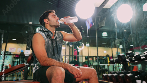 Asian male bodybuilder is sitting on fitness bench and drinking water to cool down his body after workout in gym. Shot of athlete holding a water bottle. Health care, fitness and lifestyle concept