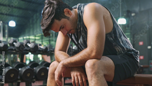 Close-up or closeup shot of Asian male bodybuilder in sportswear is sitting on fitness bench and feeling tired after weight training workout. Athlete man with exhaustion is resting on fitness bench. photo