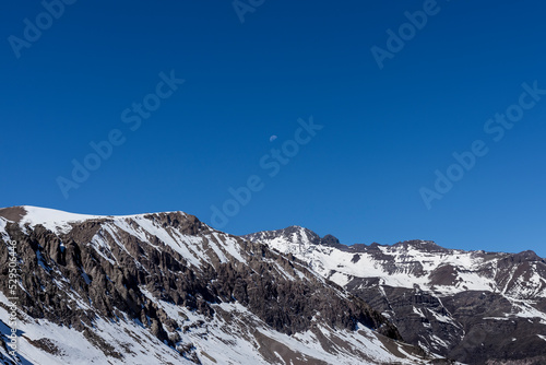 Montaña rocosa llena de nieva con cielo azul despejado y media luna
