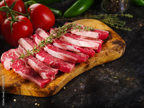 Raw meat steaks on a wooden cutting board with a sprig of rosemary, fresh tomatoes, peppers on a dark background. Cooking meat dishes. Recipes for restaurant and home cooking.