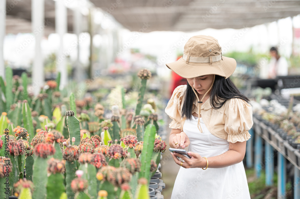 Female farmer standing holding a tablet smiling at the camera while checking quality of various cactus plants in pots with a blurred cactus in the foreground in cactus greenhouse farm.