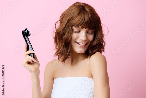 a happy, joyful, young woman stands with beautifully styled hair on a pink background and holding a black electric massager in her hand, turns her head away looking at the bottom