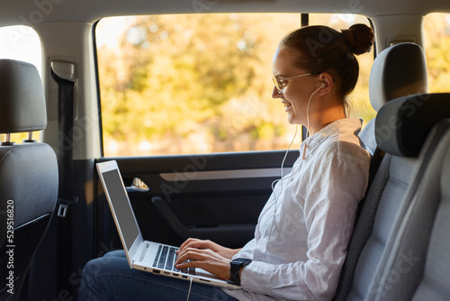 Profile portrait of smiling delighted Caucasian young business woman using portable computer while sitting in the back seat of car, typing on laptop, expressing positive emotions.