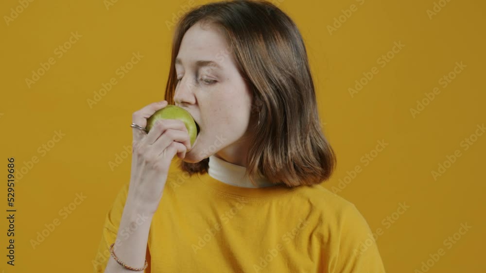 Slow motion portrait of slender young woman eating apple and smiling on yellow background. Healthy nutrition and lifestyle concept. Slow motion portrait of slender young woman eating apple and smiling on yellow background. Healthy nutrition and lifestyle concept.