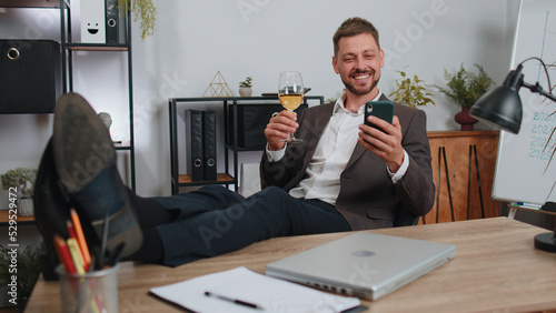 Cheers. Businessman drinking champagne from glass after working, putting legs on table sitting at office. Happy male manager freelance man celebrate successful contract agreement holding smartphone