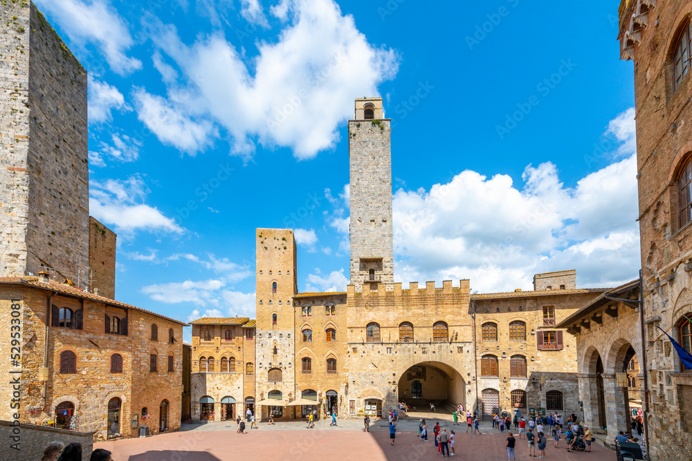 Tourists on a summer day with blue skies over the Torre Rognosa and Salvucci Towers of the Piazza Duomo in the historic Tuscan hill town of San Gimignano, Italy.