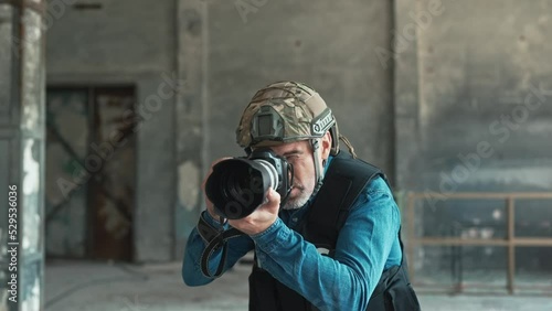 One man in helmet and bulletproof vest holding camera taking photos in war zone. Male war journalist with digital camera at the place of action in damaged building working. War press. Armed conflict photo