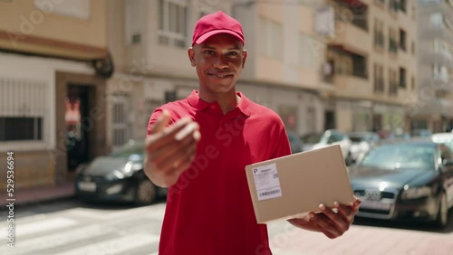 Young latin man delivery worker holding package doig coming gesture with hand at street photo