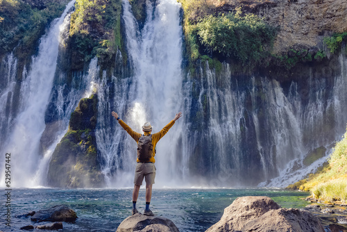 Man traveler standing with his hands raised with a view on the beautiful waterfall, view from the back