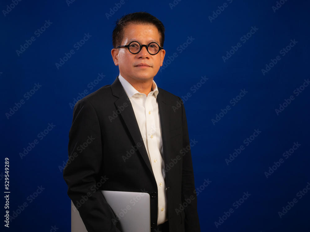 Portrait of smart middle-aged asian confident successful businessman wearing suit holding laptop isolated on blue background in studio, looking attractive and positive professional leadership Portrait of smart middle-aged asian confident successful businessman wearing suit holding laptop isolated on blue background in studio, looking attractive and positive professional leadership