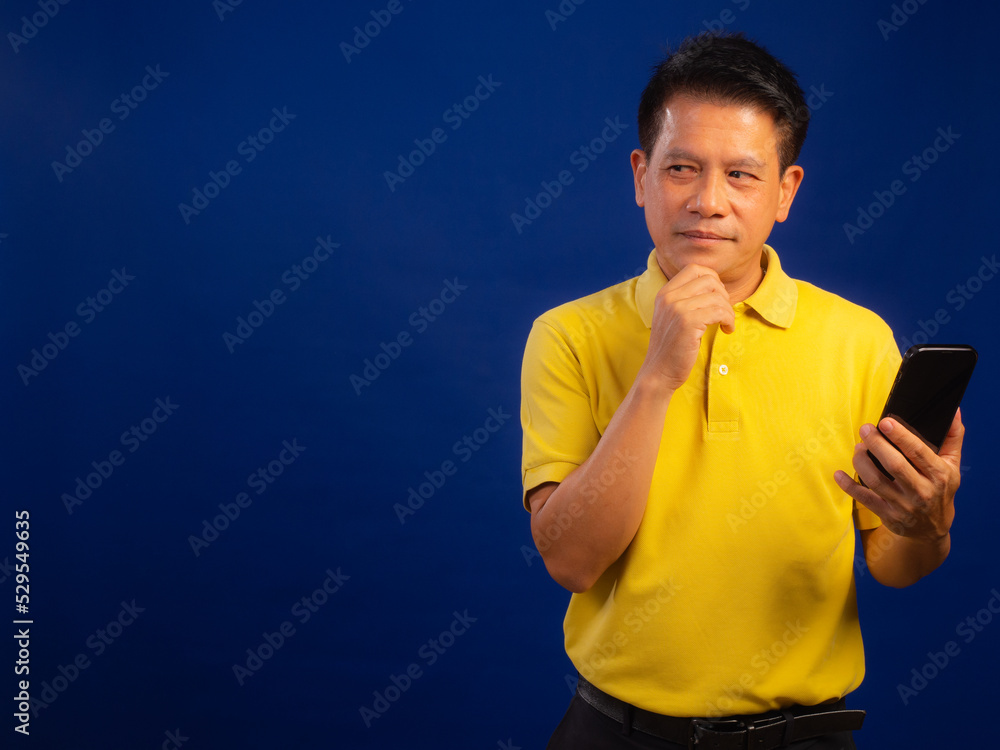 Portrait of smart middle-aged asian man in casual yellow T-shirt holding smartphone isolated on blue background with copyspace in studio, looking attractive Portrait of smart middle-aged asian man in casual yellow T-shirt holding smartphone isolated on blue background with copyspace in studio, looking attractive