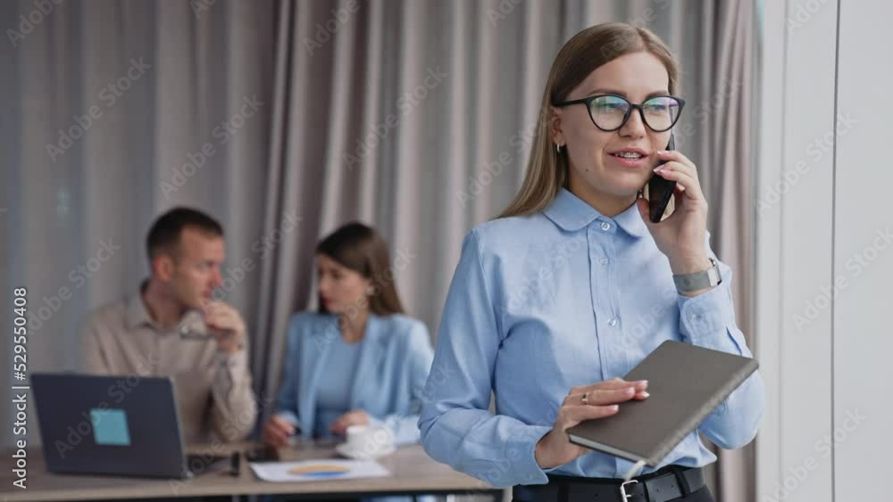 Phone conversation in the office near window. Lady in glasses speaking on the phone holding paper notebook in her hand. Colleagues at desk at backdrop. Phone conversation in the office near window. Lady in glasses speaking on the phone holding paper notebook in her hand. Colleagues at desk at backdrop.