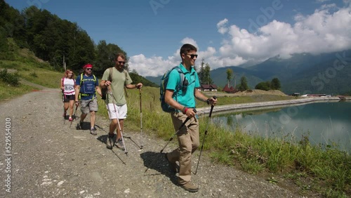 Group of young hikers walks down past transparent blue mountain lake. Instructor confidently leads group to goal. The fight against hypodynamia for white collars photo