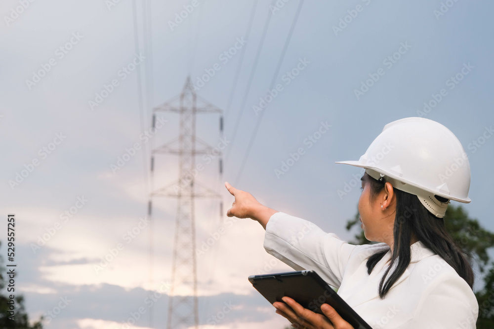 Asian female electrical engineer working on laptop near high voltage pole, inspecting power grid.