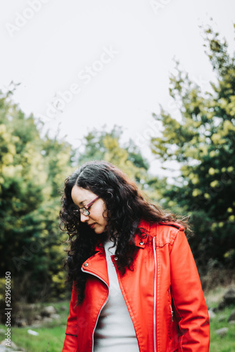 curly brunette latin woman looking down, wearing a red leather jacket in nature.