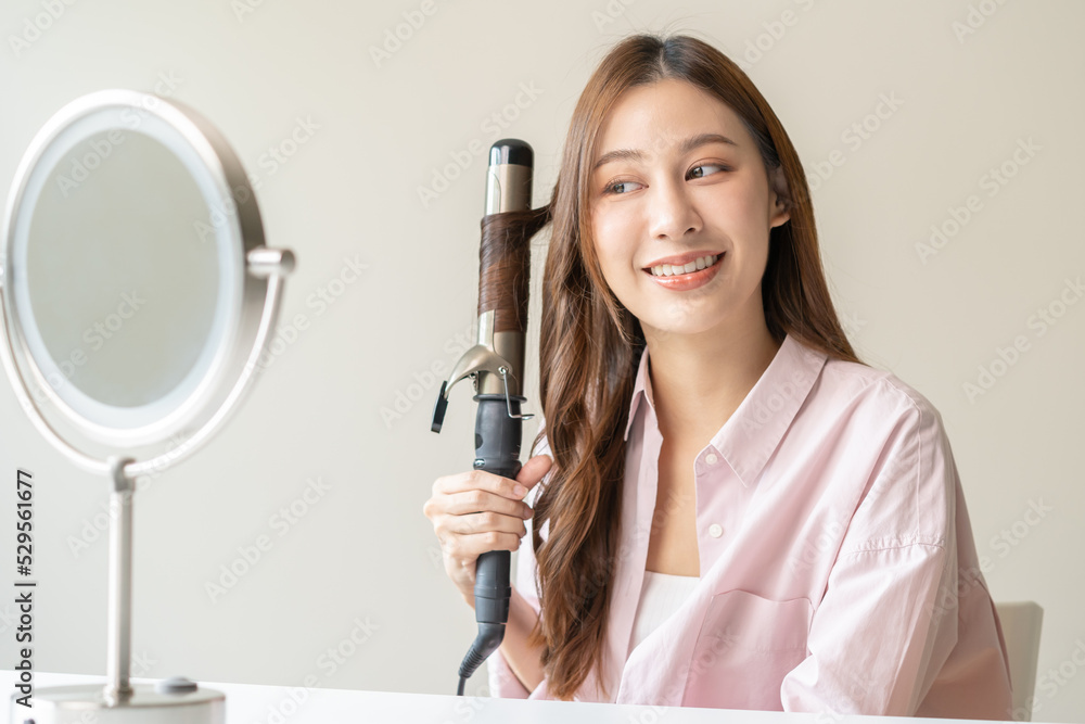Beautiful curly brunette, attractive asian young woman, girl smiling using curler, curling iron long hair with ceramic curler straightener at home. Hairstyle and Hairdressing. Beauty and hair care . Beautiful curly brunette, attractive asian young woman, girl smiling using curler, curling iron long hair with ceramic curler straightener at home. Hairstyle and Hairdressing. Beauty and hair care .