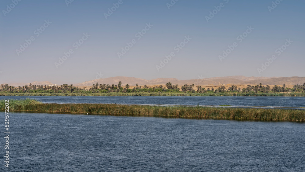 A grassy island is visible in the bed of the blue river. There is green vegetation on the shore. Sand dunes against a clear sky. Egypt. Nile