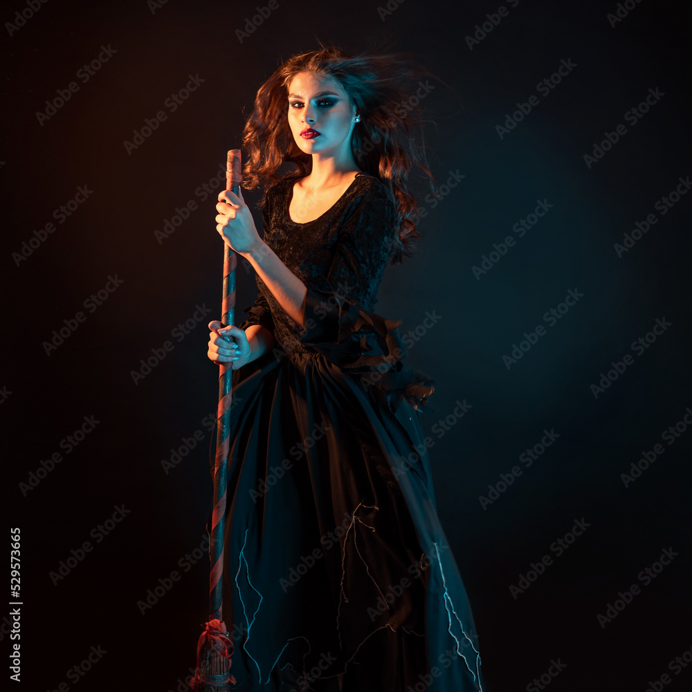 A young witch is rushing to the sabbath. A young beautiful brunette in a witch dress is standing with a broom in her hands, her hair is fluttering in the wind, warm and cold figure illumination,