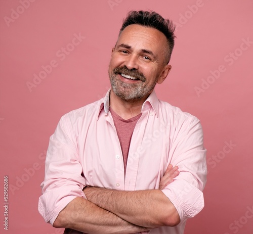 Smiling man posing with arms crossed on pink background. Portrait of attractive handsome mature man in rose shirt with crossed arms. Healthy happy smile senior beard caucasian man isolated on pink.