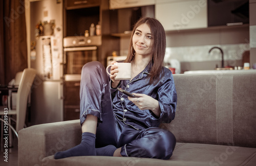 Young woman in nightie drinks coffee and watches TV while sitting on the sofa in the living room