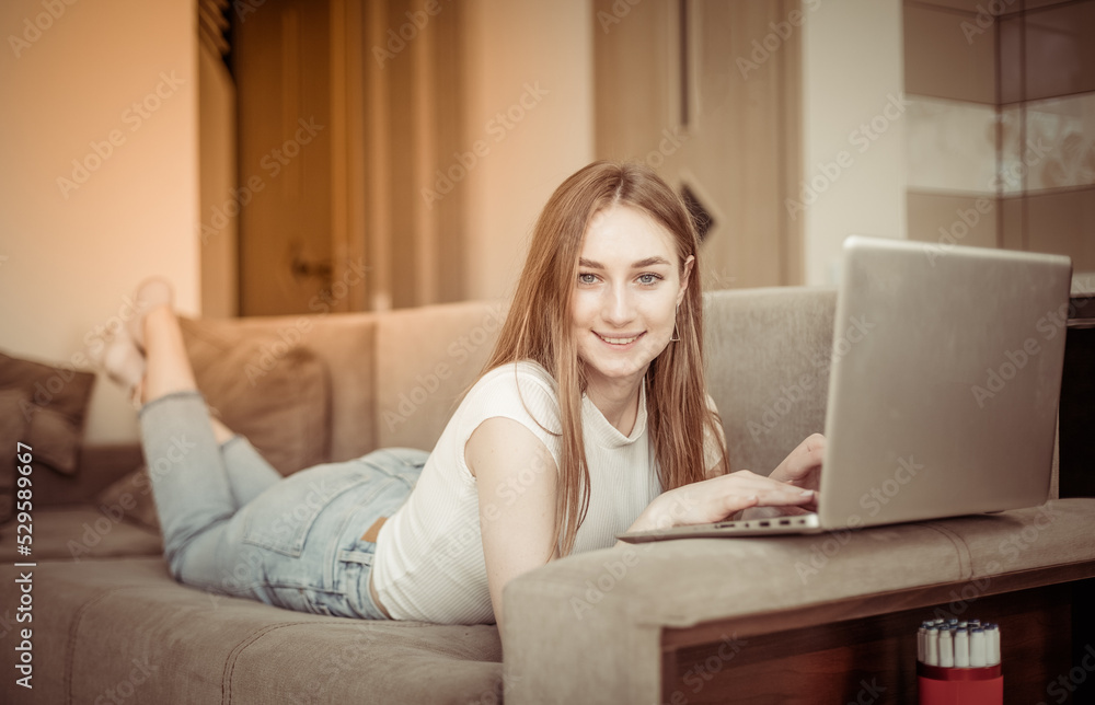 Young cute smiling woman with laptop looking at camera while lies on sofa in living room Young cute smiling woman with laptop looking at camera while lies on sofa in living room
