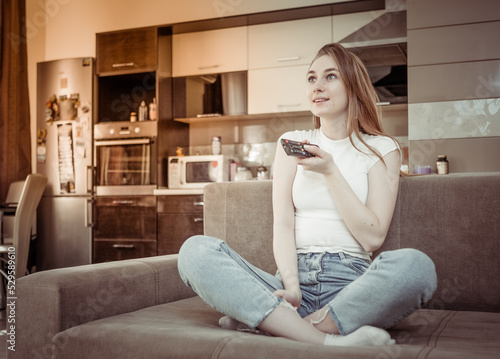 Happy woman looks at TV screen and switches TV channels with a remote control, sitting on sofa in living room. © splitov27