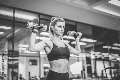 Sexy young fit woman with perfect body dressed in sportswear trains shoulder muscles with dumbbells in modern gym. Healthy lifestyle