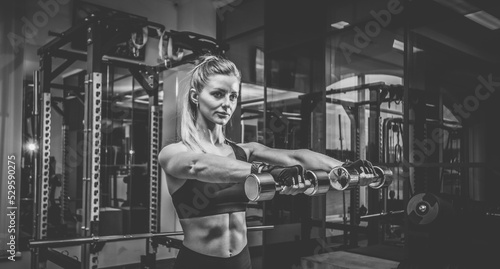 Sexy young fit woman with perfect body dressed in sportswear trains shoulder muscles with dumbbells in dark gym. Healthy lifestyle
