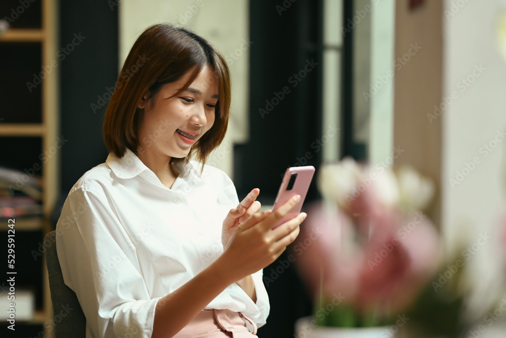 Smiling woman employee sitting front of laptop at her office desk and using mobile phone Smiling woman employee sitting front of laptop at her office desk and using mobile phone